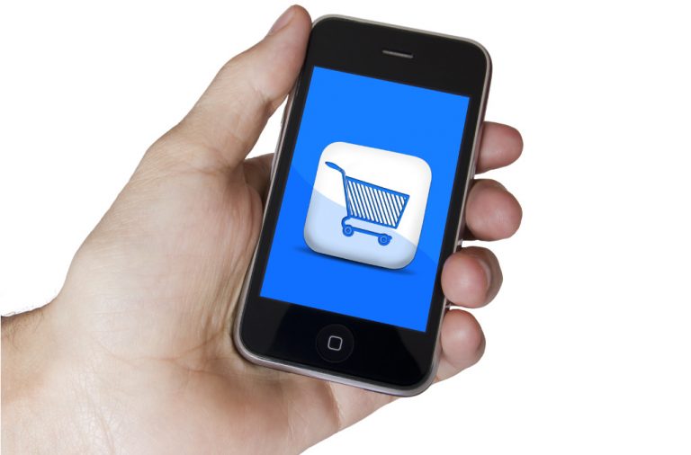 A mão de um pessoa segurando um celular e na tela do celular aparece um carrinho de compras, demonstrando o e-commerce mobile