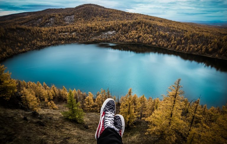 Um lago envolto de uma linda vegetação e na parte baixa da imagem, aparecem dois pés cruzados, sugerindo que alguém está tranquilamente descansando e admirando a paisagem