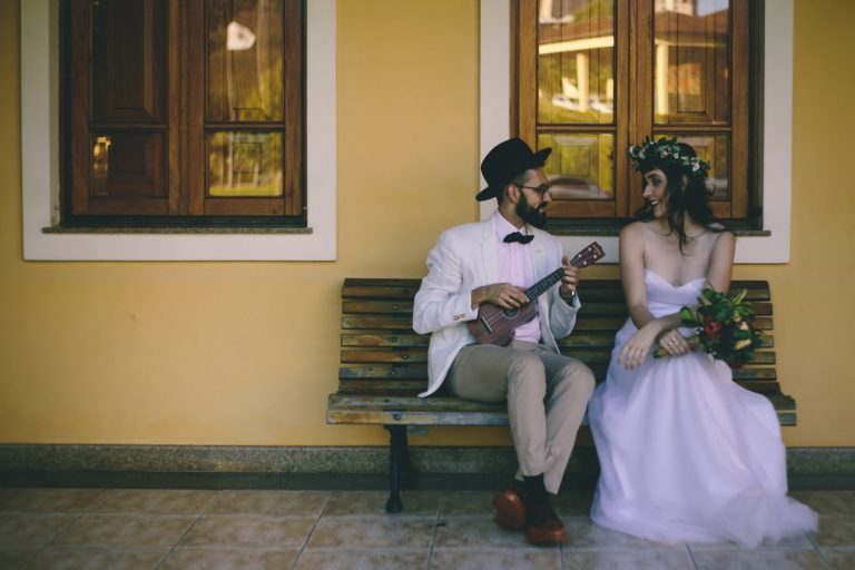 Casal sentado em um banco, ela de vestido de noiva e ele de roupa esporte-social, representando um ecommerce de vestidos de noiva despojados